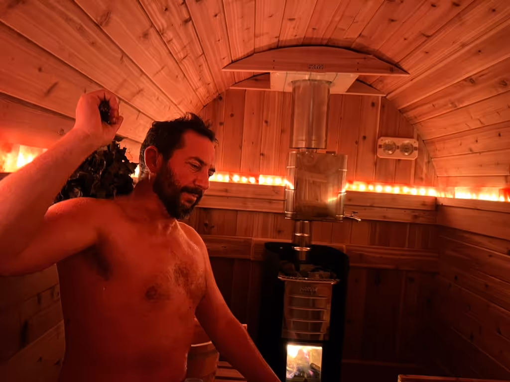Two men sit and relax inside the warm, ambient light of a wooden barrel sauna.
