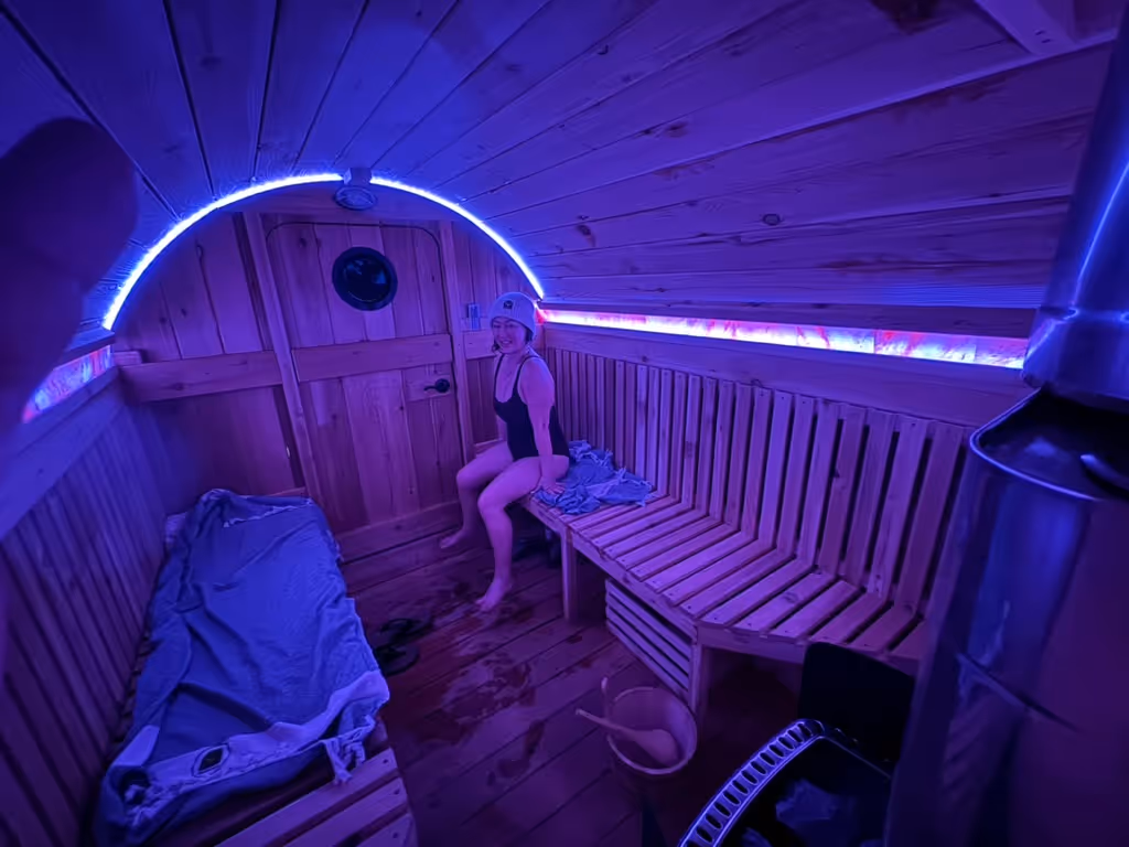 A person relaxes on a bench inside a barrel sauna that is lit with cool blue LED light.