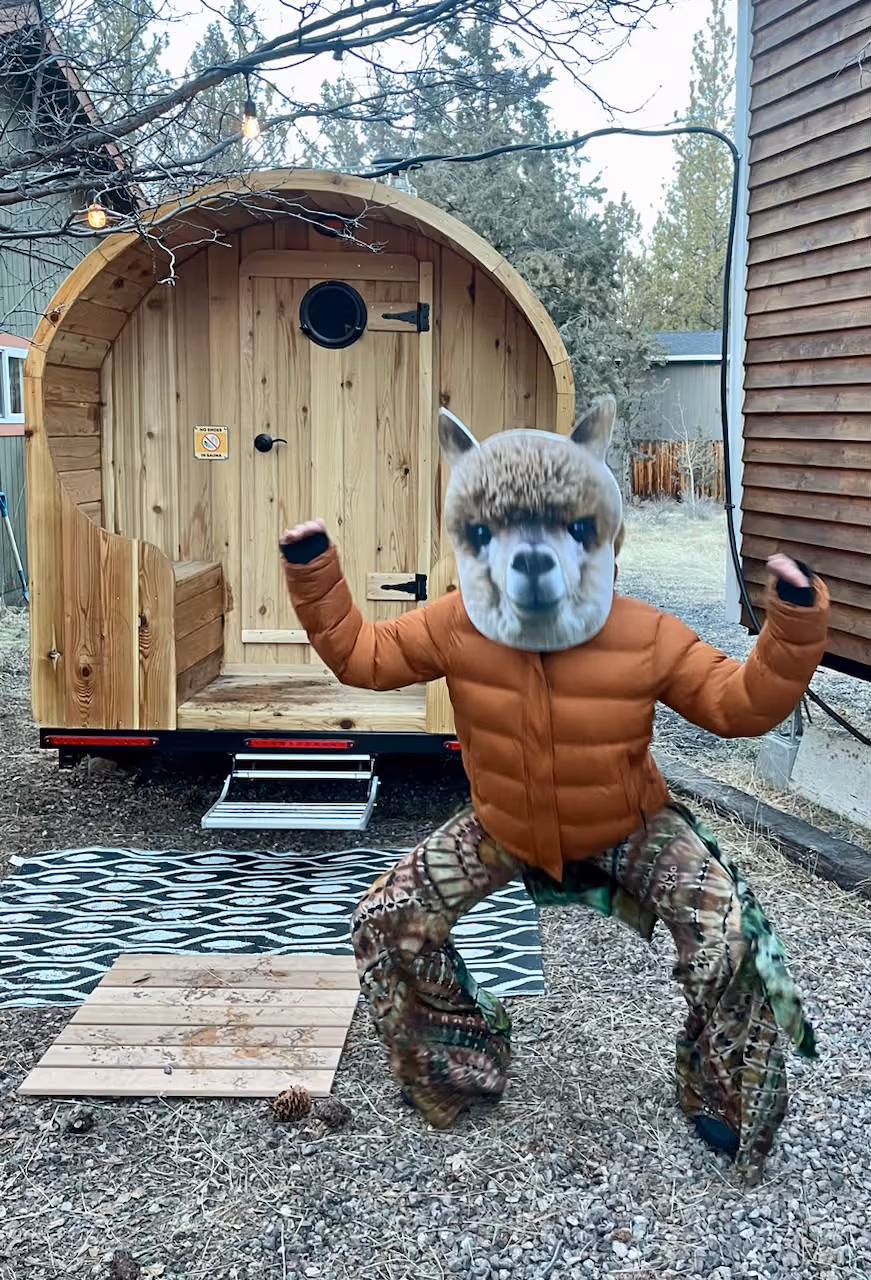 A muscular man wearing a winter hat flexes his biceps while standing in the doorway of a mobile sauna.