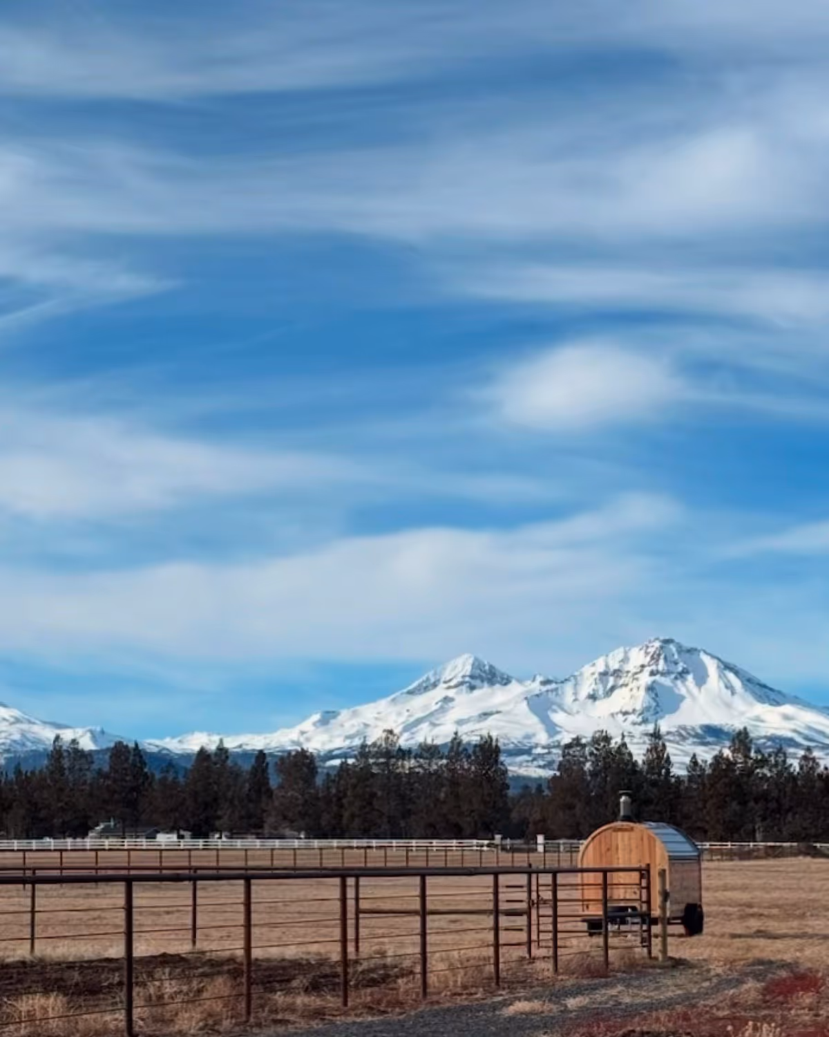 A mobile barrel sauna positioned in a field with a large, snow-capped mountain featured prominently in the background.