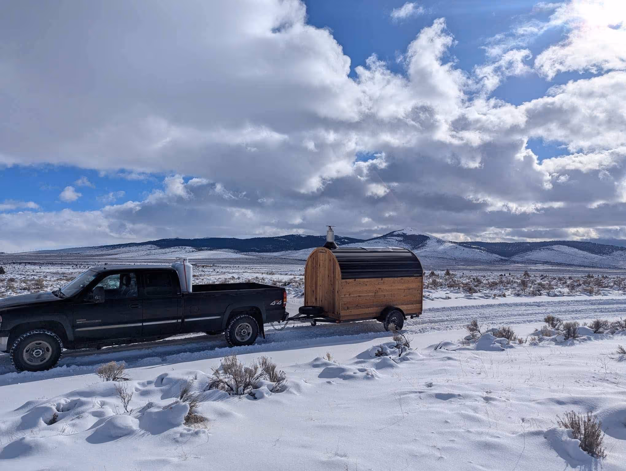Two mobile barrel saunas are parked in a wide, snow-dusted landscape under a cloudy sky.