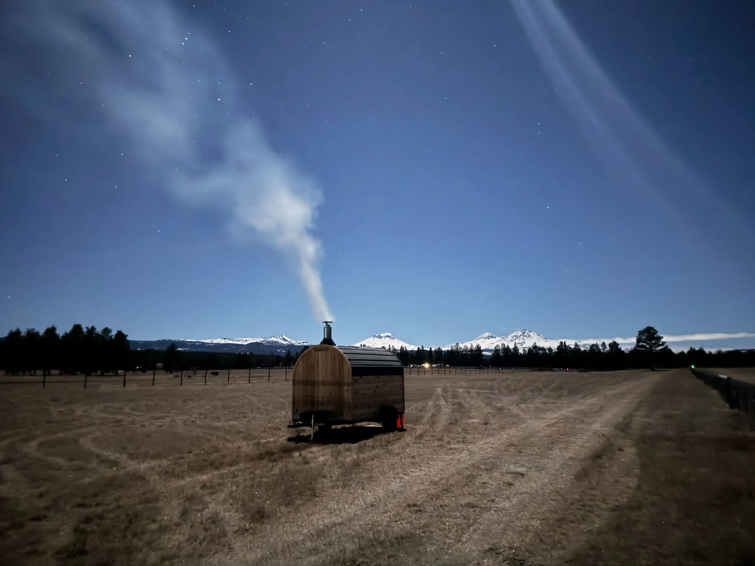 A lone mobile sauna sits in a vast, dry field, with steam rising from its chimney into a blue sky.