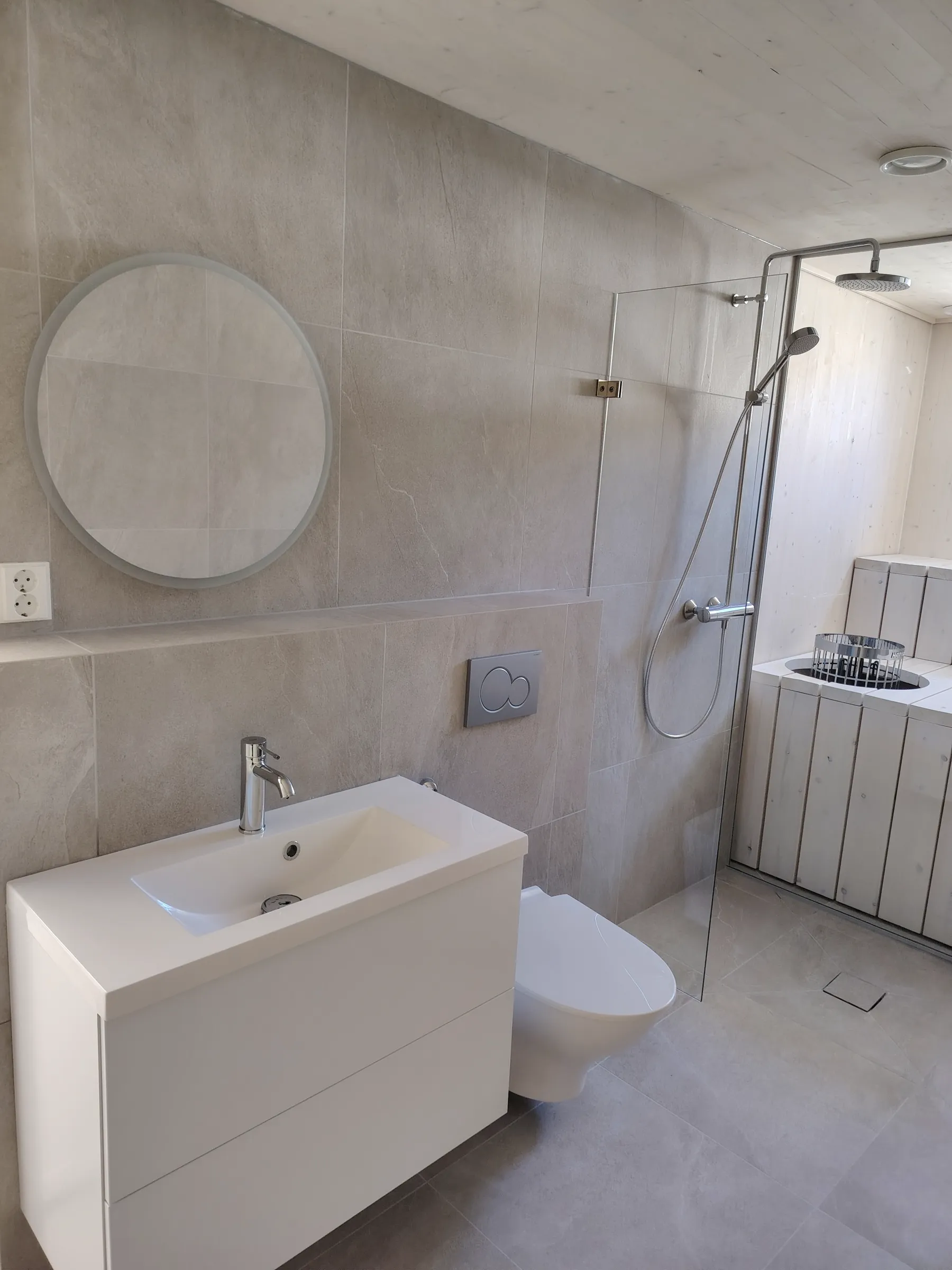 Moderni kylpyhuone, jossa on pyöreä peili, valkoinen pesuallas, seinä-wc ja suihkukaappi, jossa on puinen sauna. Seinät on laatoitettu harmaalla laatalla.