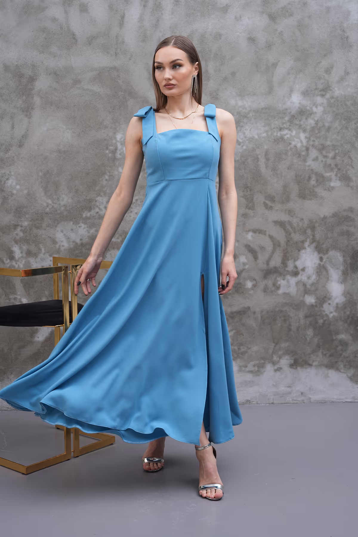 Model wearing the Celina sky blue A-line dress with shoulder bows and front slit, twirling gracefully in front of a textured wall.