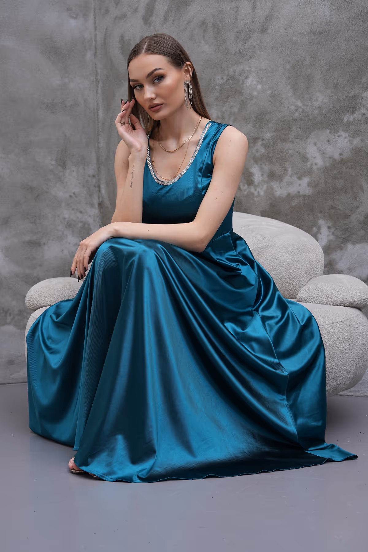 Model sitting on a beige chair in a teal Liona satin gown, showcasing the dress's full-length flow and subtle shimmer.