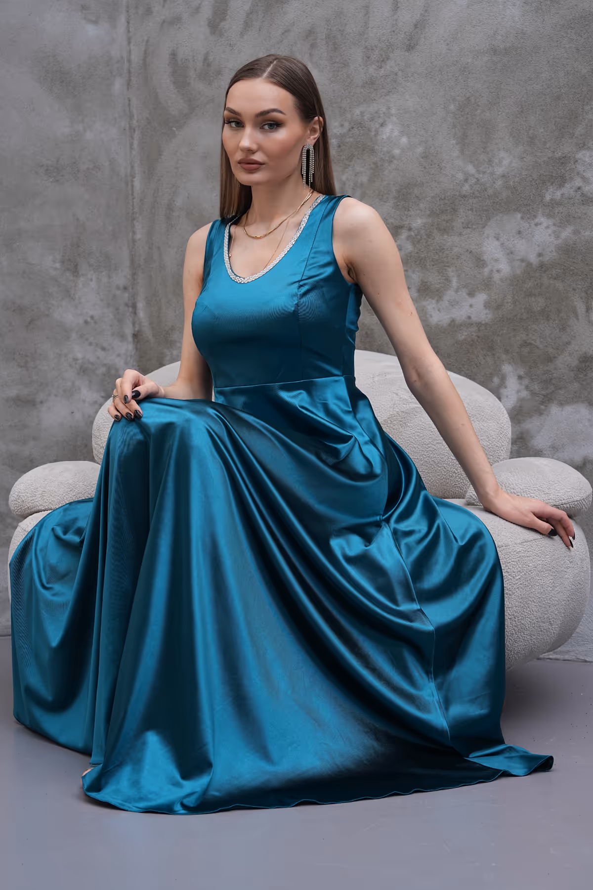 Seated pose highlighting the glossy texture and elegant drape of the Liona teal gown with a sleeveless bodice.