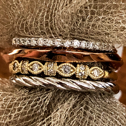 Stack of mixed-metal rings including pavé diamond, hammered rose gold, milgrain yellow gold, and twisted cable bands, styled on textured fabric