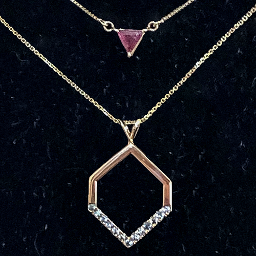 Layered gold necklaces featuring a small triangle-cut pink stone pendant above a geometric gold pendant with diamond accents