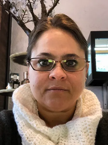 Donna, CAD designer at Esch Metals Studio, wearing glasses and a white knit scarf inside the shop