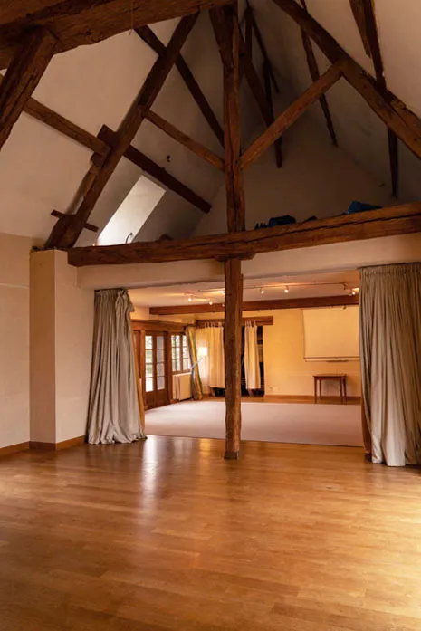 Grande salle vide avec plancher en bois, poutres apparentes en bois foncé et rideaux beige.