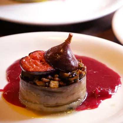 Plat gastronomique avec une tranche de figue, un figue entier et des noix sur un lit de sauce rouge.
