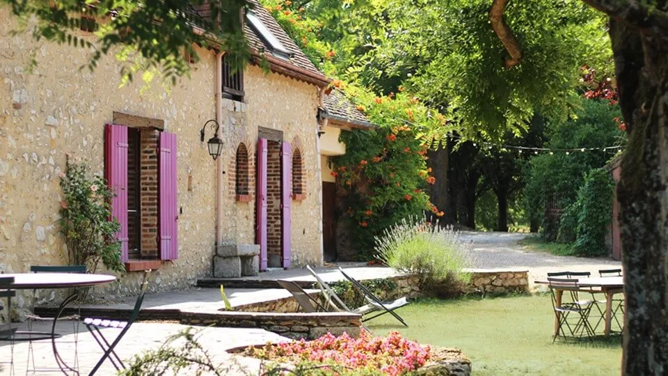 Façade d'une maison en pierre avec des volets violets entourée d'un jardin verdoyant avec des tables de jardin et des chaises sous des arbres.