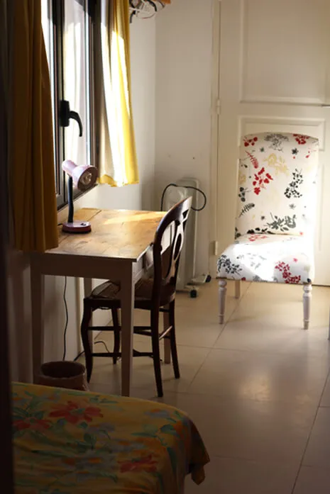 Une pièce avec un bureau en bois clair près d'une fenêtre avec des rideaux jaunes, une lampe de bureau rose, une chaise en bois et un fauteuil à motifs floraux éclairé par la lumière naturelle.