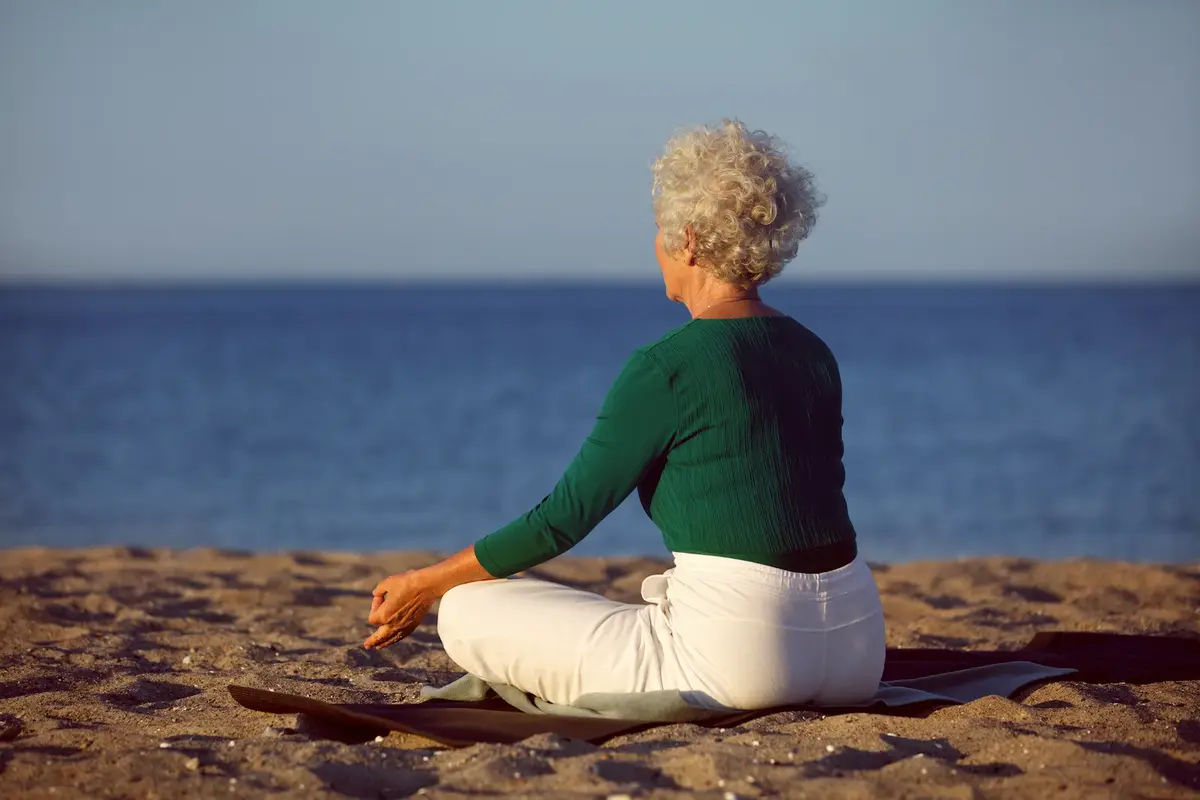 Une femme aux cheveux gris, assise de dos, regarde la mer, en méditant.