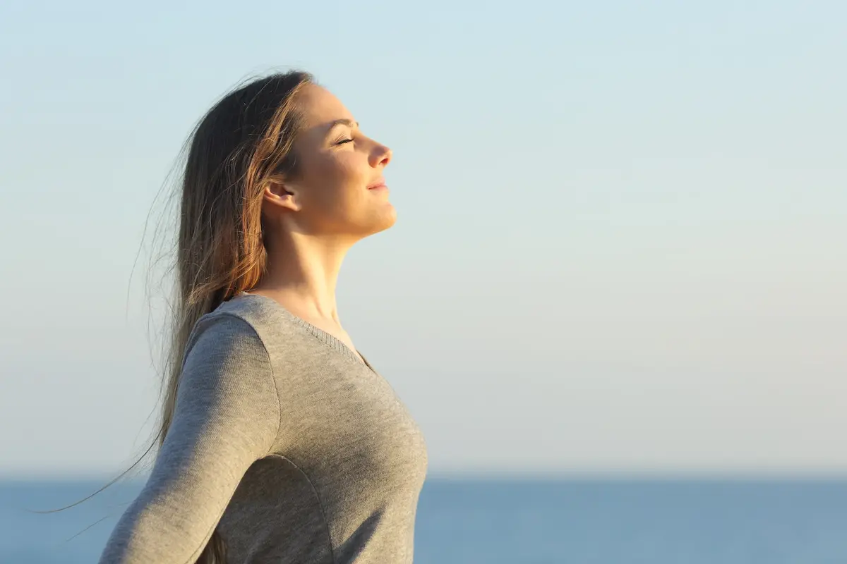 Une jeune femme souriante respire profondément face à la mer.