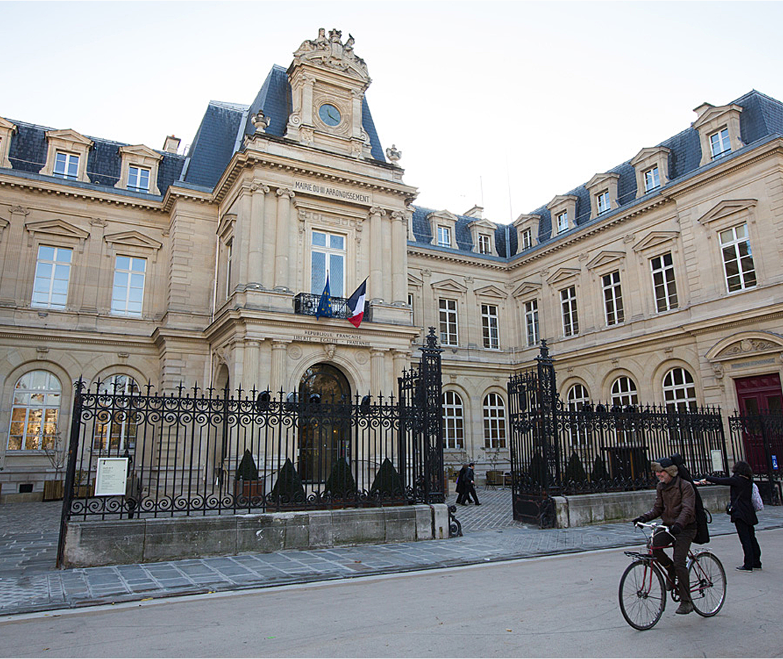 Façade historique de la mairie du 11e arrondissement de Paris avec un portail en fer forgé, deux drapeaux et un homme à vélo au premier plan.