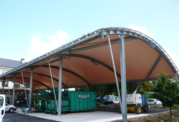 Abri avec une structure de toit incurvée soutenue par des piliers métalliques couvrant des équipements industriels verts.