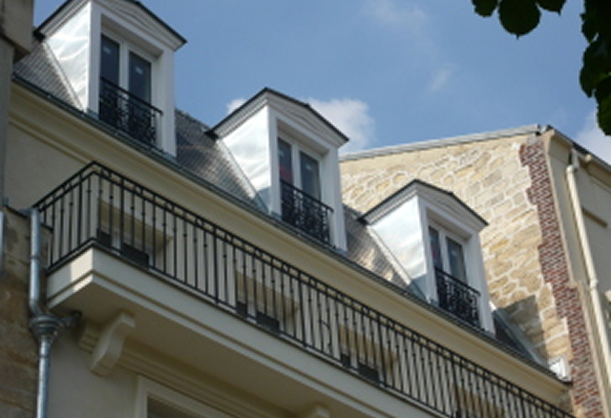 Façade d'un bâtiment avec trois lucarnes blanches et un balcon en fer forgé sous un ciel bleu.