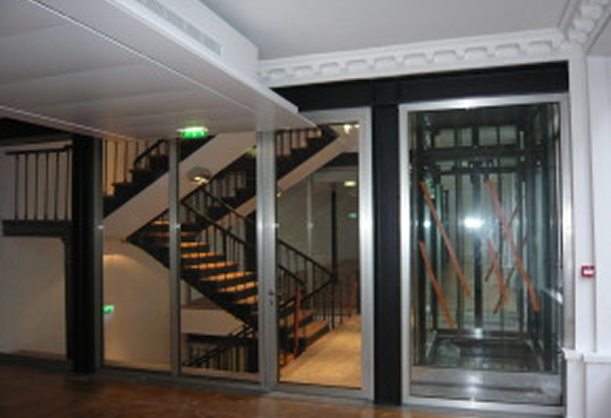 Intérieur d'un bâtiment avec un escalier en métal visible derrière une cloison vitrée et un ascenseur équipé de renforts en bois.