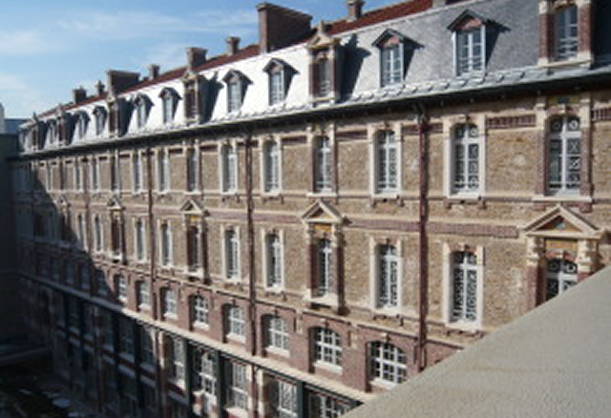 Façade d'un grand bâtiment ancien en briques avec de nombreuses fenêtres et un toit à lucarnes.