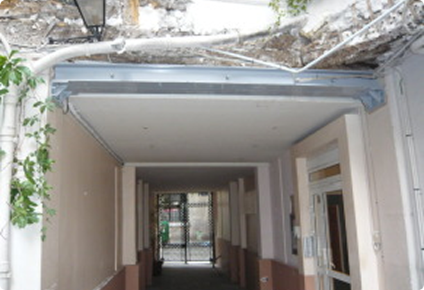 Passage couvert d'immeuble avec une structure métallique renforcée fixée au plafond et un mur en pierre ancien au-dessus.