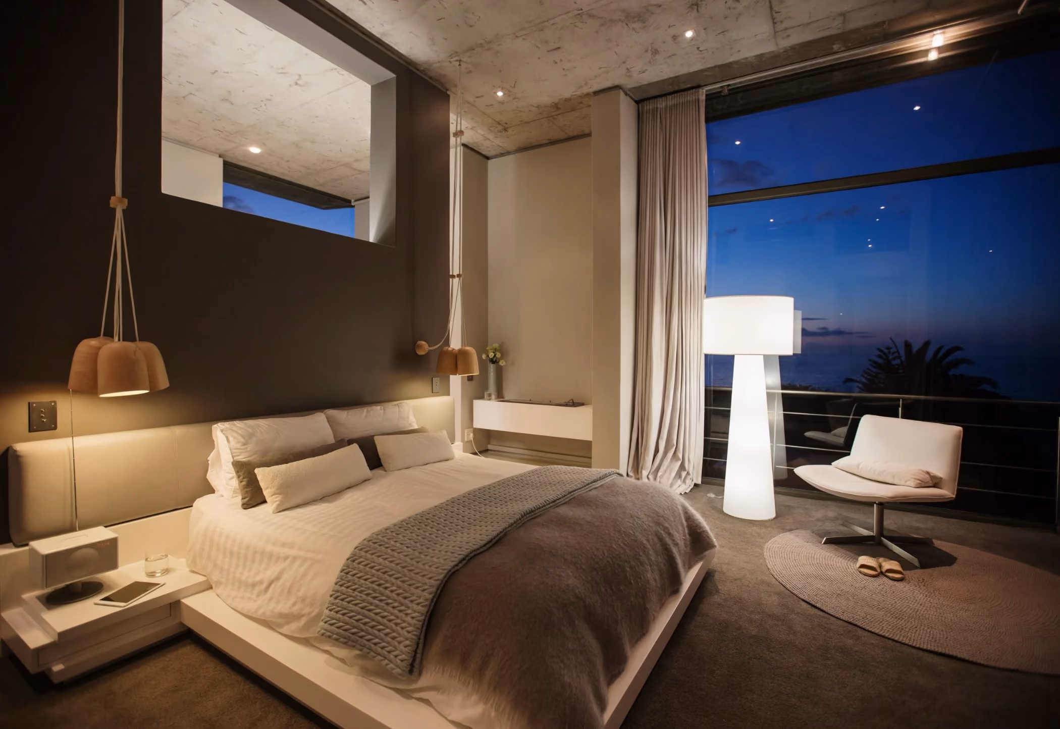 Modern bedroom with a large bed, neutral bedding, pendant lights, and a white chair beside a large window showing a night sky.
