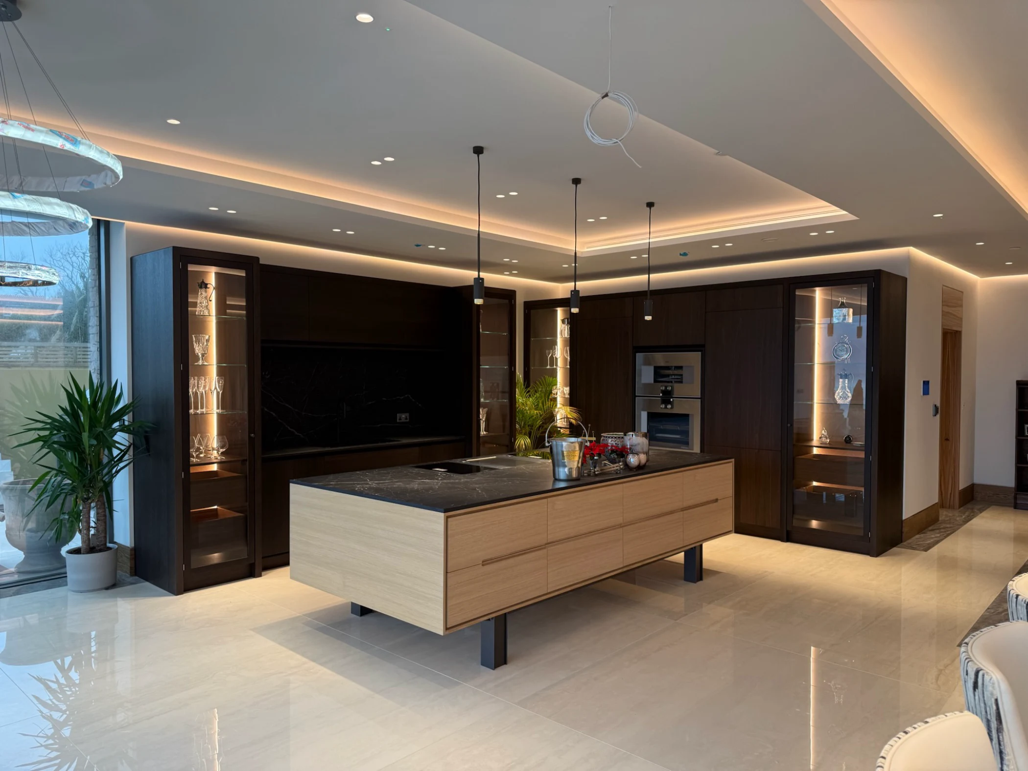 Modern kitchen with a large wooden island featuring a black marble countertop, dark wood cabinets, glass display cabinets with lighting, and pendant lights hanging from the ceiling.