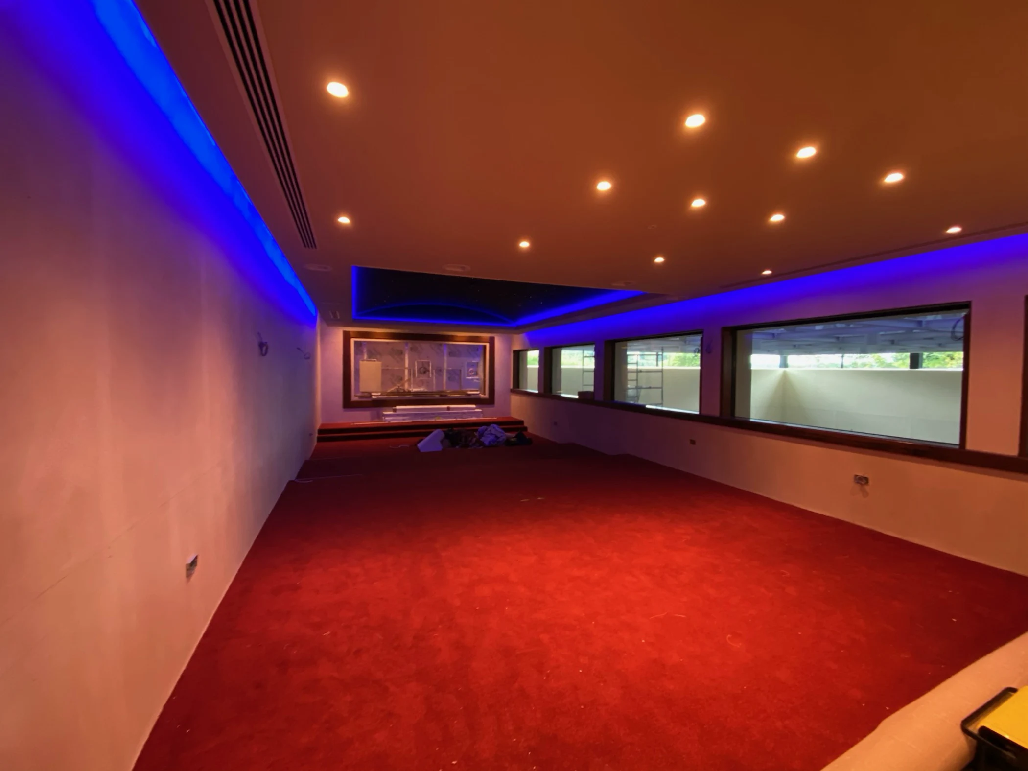 Spacious room with red carpet, recessed ceiling lights, blue LED strip lighting, and large windows along one wall.