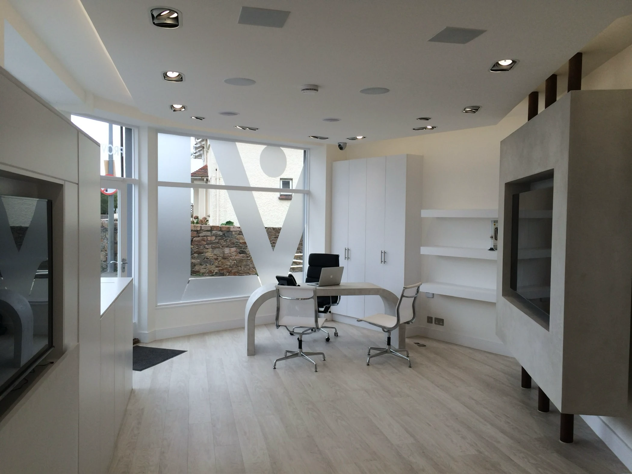 Modern office with curved desk, two office chairs, a black chair, laptop, large frosted window, white cabinets, shelves, and light wood flooring.