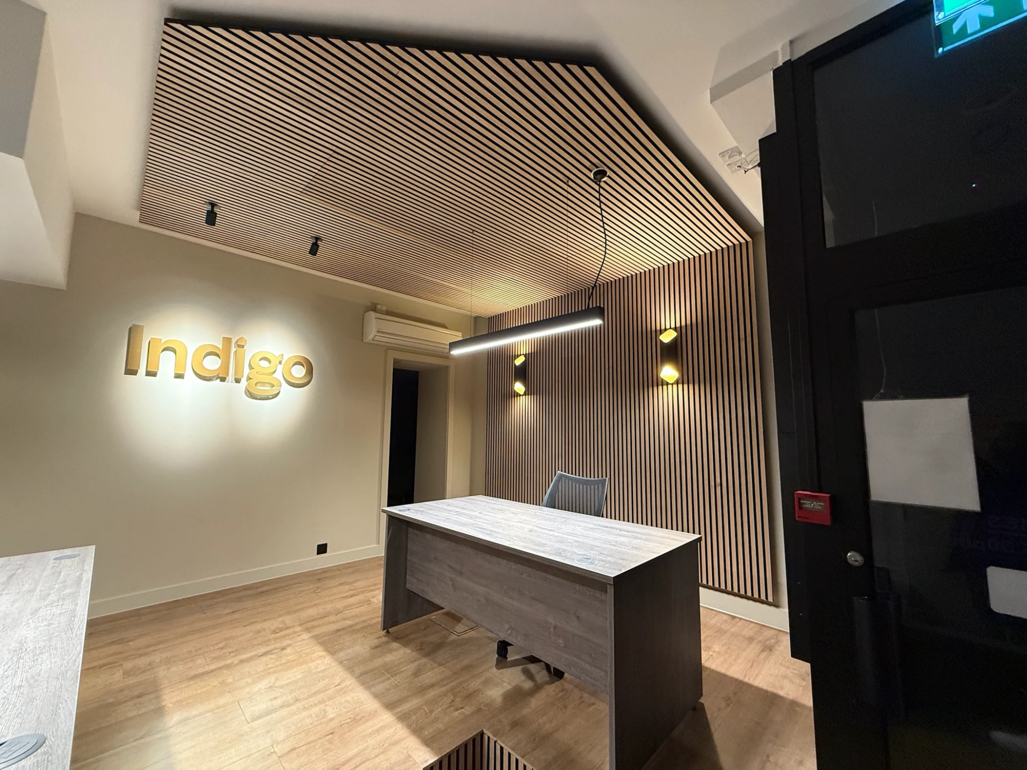 Modern office reception with wooden flooring, a desk, a gray chair, vertical wood panel walls, and illuminated 'Indigo' wall sign.