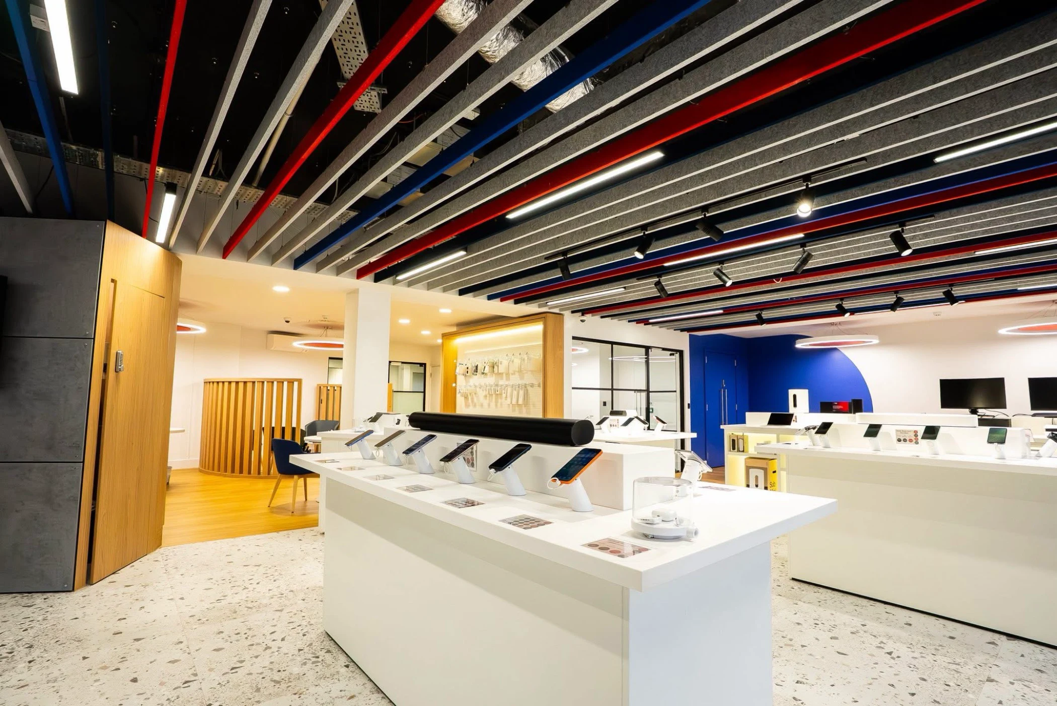 Modern electronics store interior with white display tables showcasing smartphones and accessories, colorful ceiling beams, and a blue accent wall.