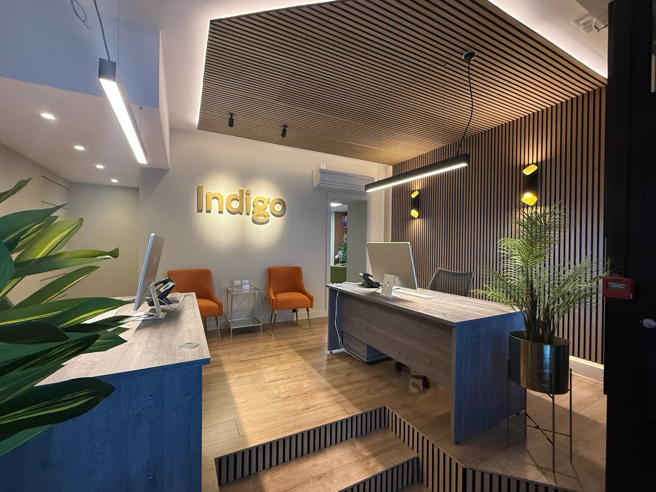 Modern office reception with wooden slatted ceiling and wall, two orange chairs, desk with computer, and indoor plants.