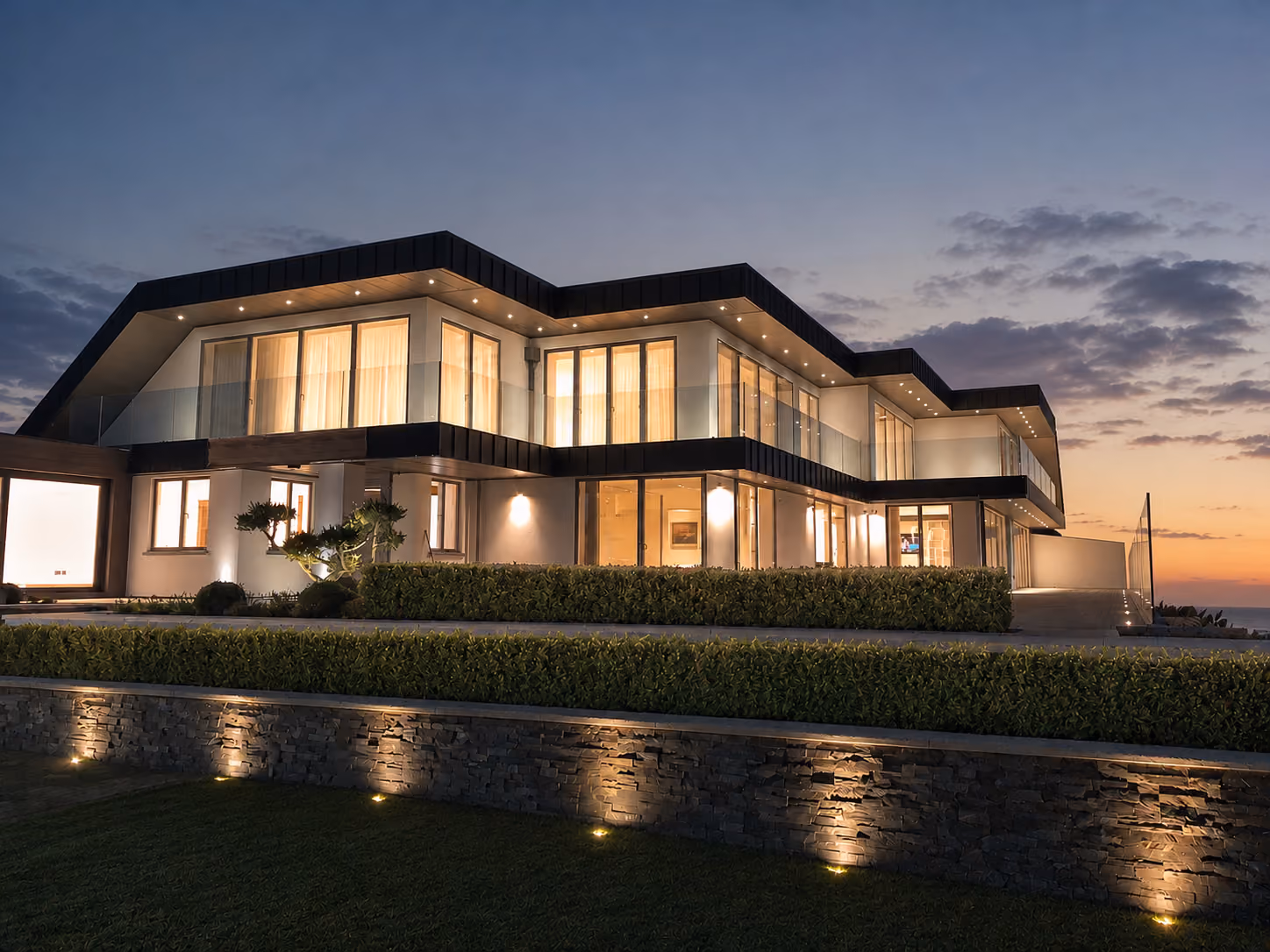 Modern two-story house at dusk with large illuminated windows, surrounded by neatly trimmed hedges and outdoor wall lighting against a cloudy sunset sky.