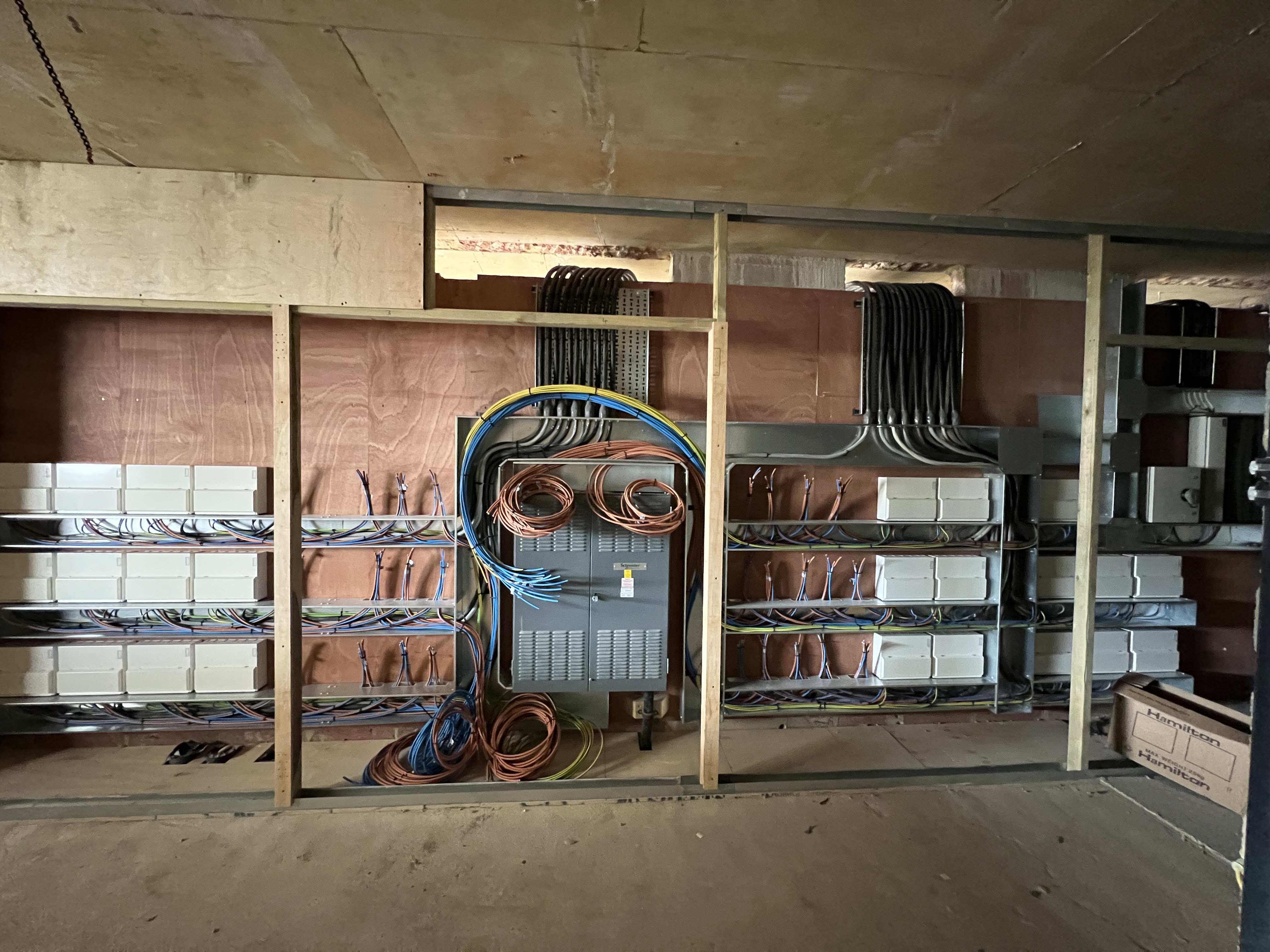 Electrical panel mounted on a plywood wall with organized wiring conduit and multiple cables coiled and running horizontally and vertically inside a room under construction or renovation.