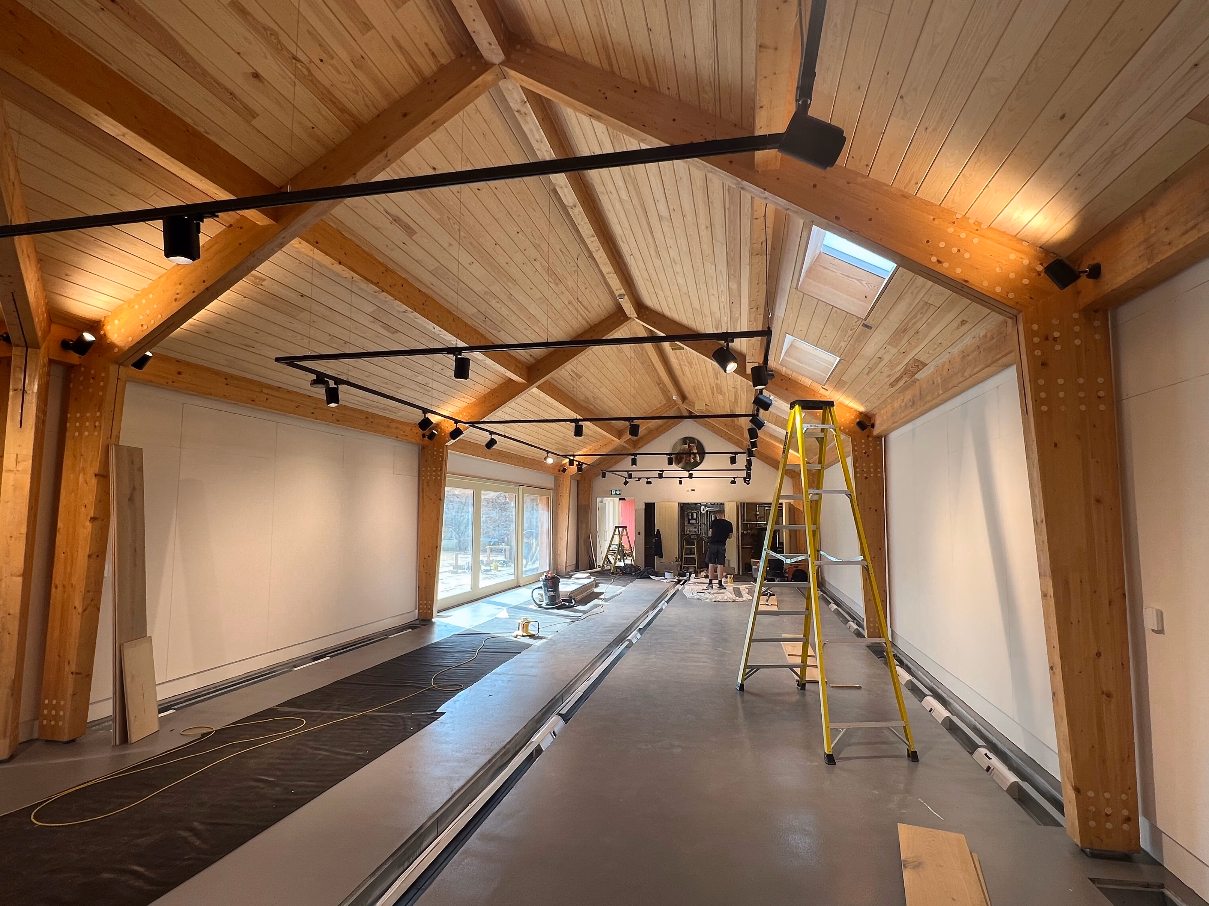 Interior of a large room with wooden beams and a wood-paneled ceiling, featuring multiple track lights, skylights, and large windows on one side, with ladders and construction materials on the floor indicating ongoing work.