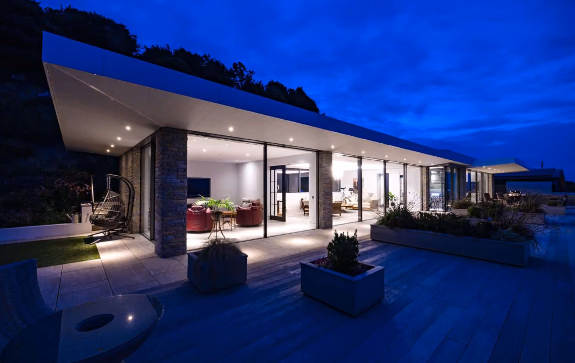 Modern two-story house with large glass windows, outdoor seating, and illuminated swimming pool at dusk.