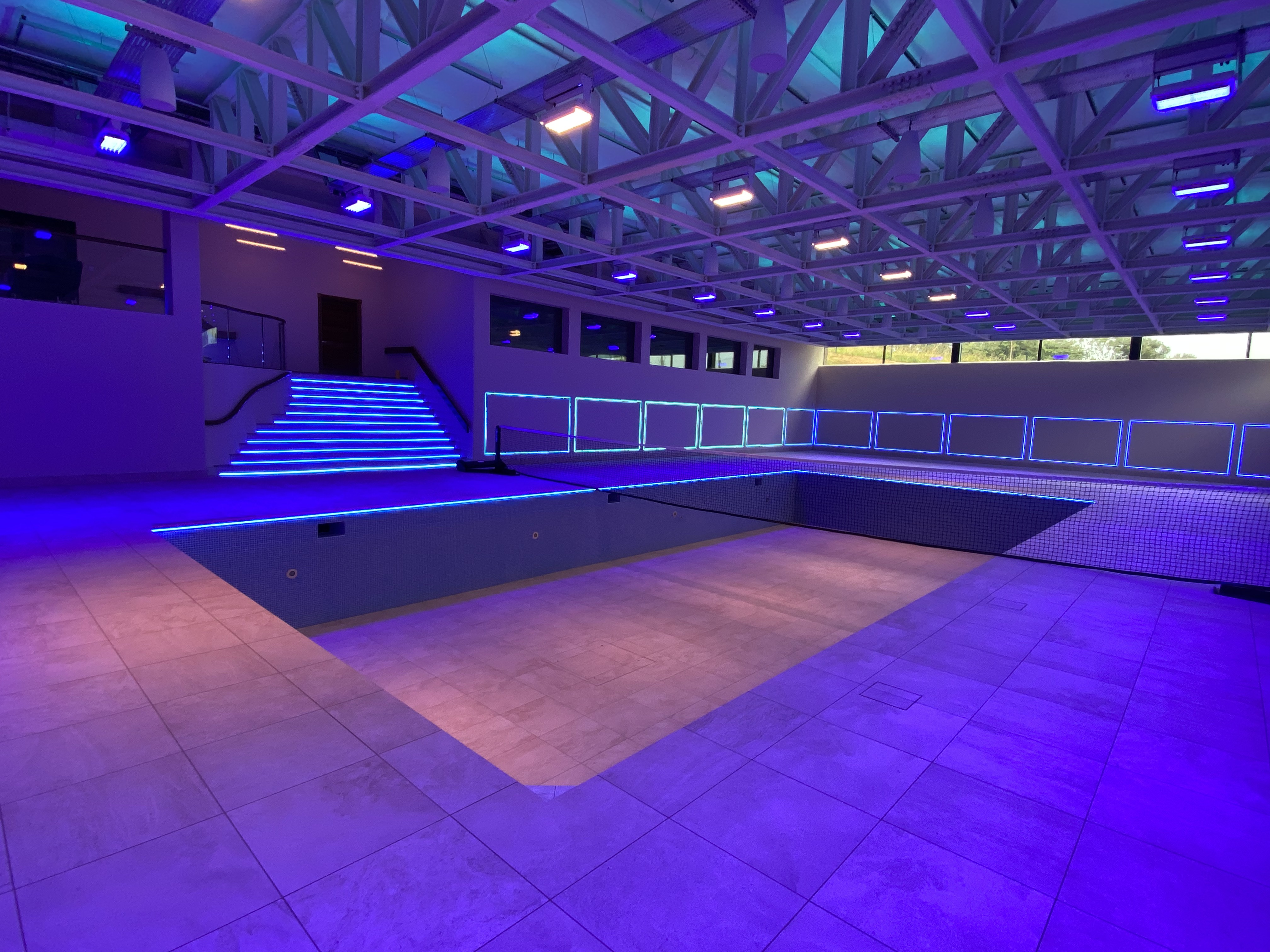 Indoor empty swimming pool with tiled floor and steps illuminated by blue LED strip lights, under a ceiling with exposed white beams and blue spotlights, large windows on one side.