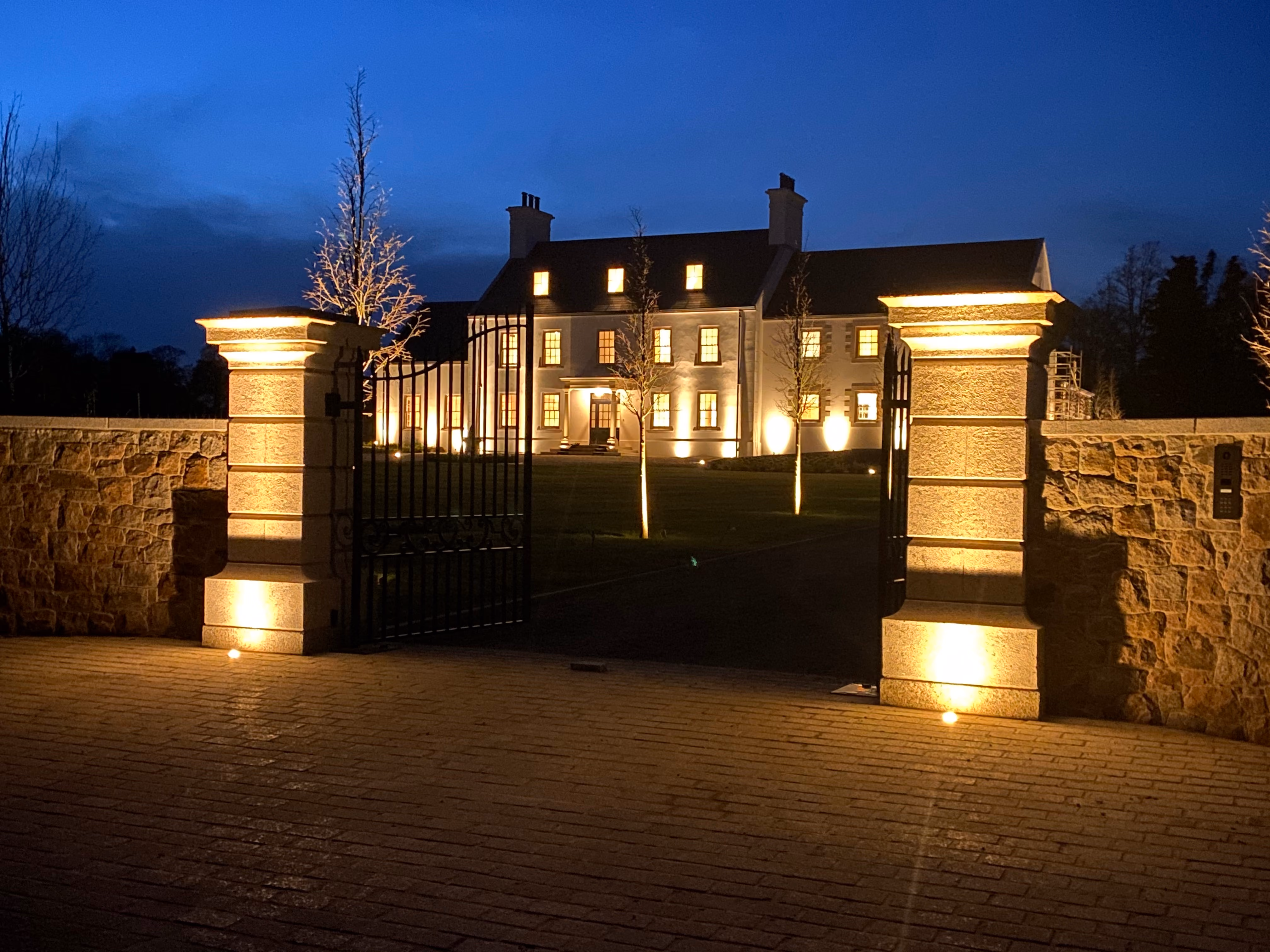 Night view of a large illuminated two-story house with multiple lit windows, visible through an open wrought iron gate set between stone pillars and stone walls, with small trees and pathway lights leading to the house.