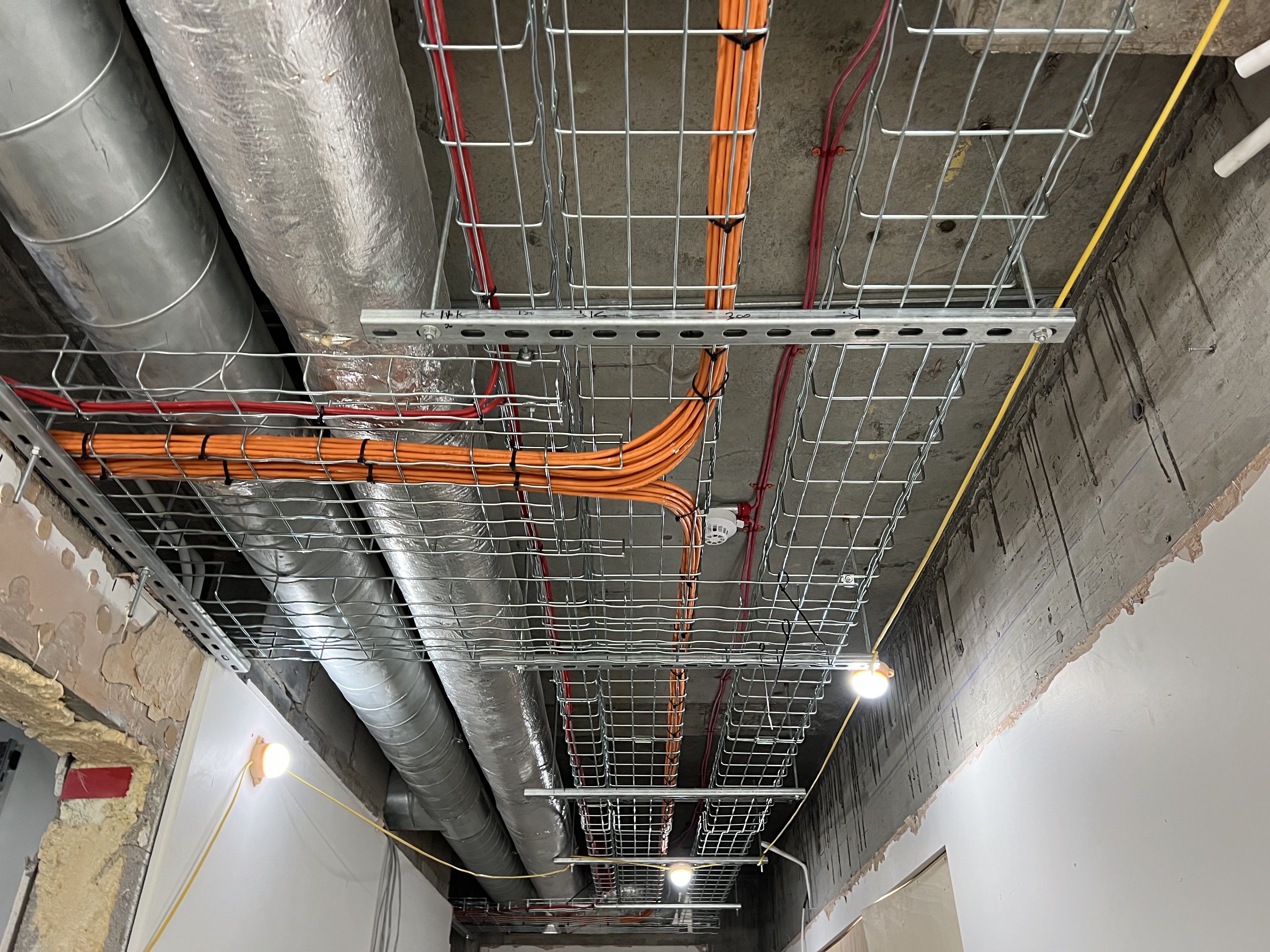 Ceiling with exposed ventilation ducts wrapped in reflective insulation, metal wire cable trays holding organized orange and red cables, and hanging temporary lights in a building under construction.