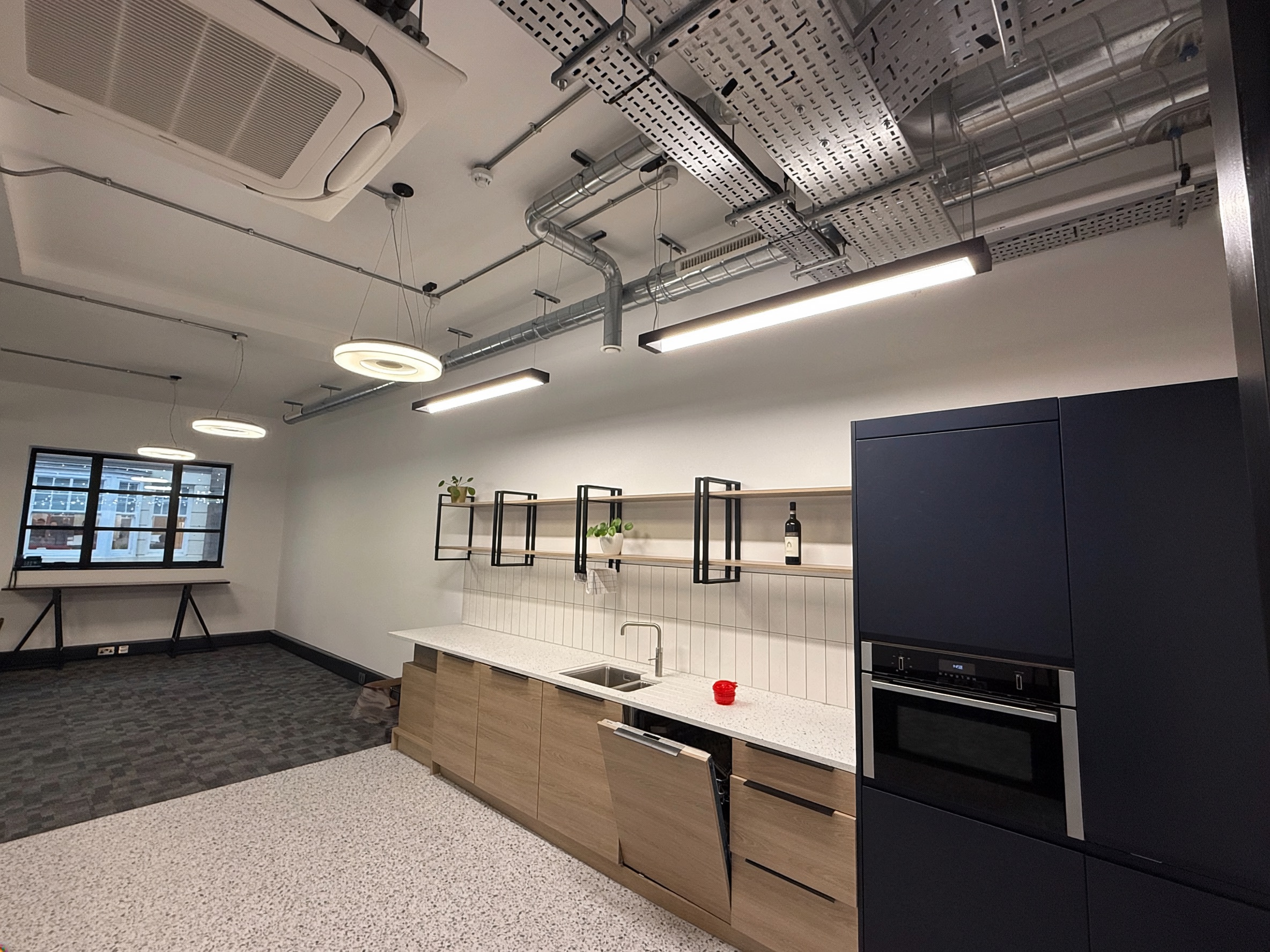 Modern kitchen area in an office with wood lower cabinets, white countertop, sink, open dishwasher, black built-in oven, open shelving with plants and wine bottle, and overhead round and linear ceiling lights.