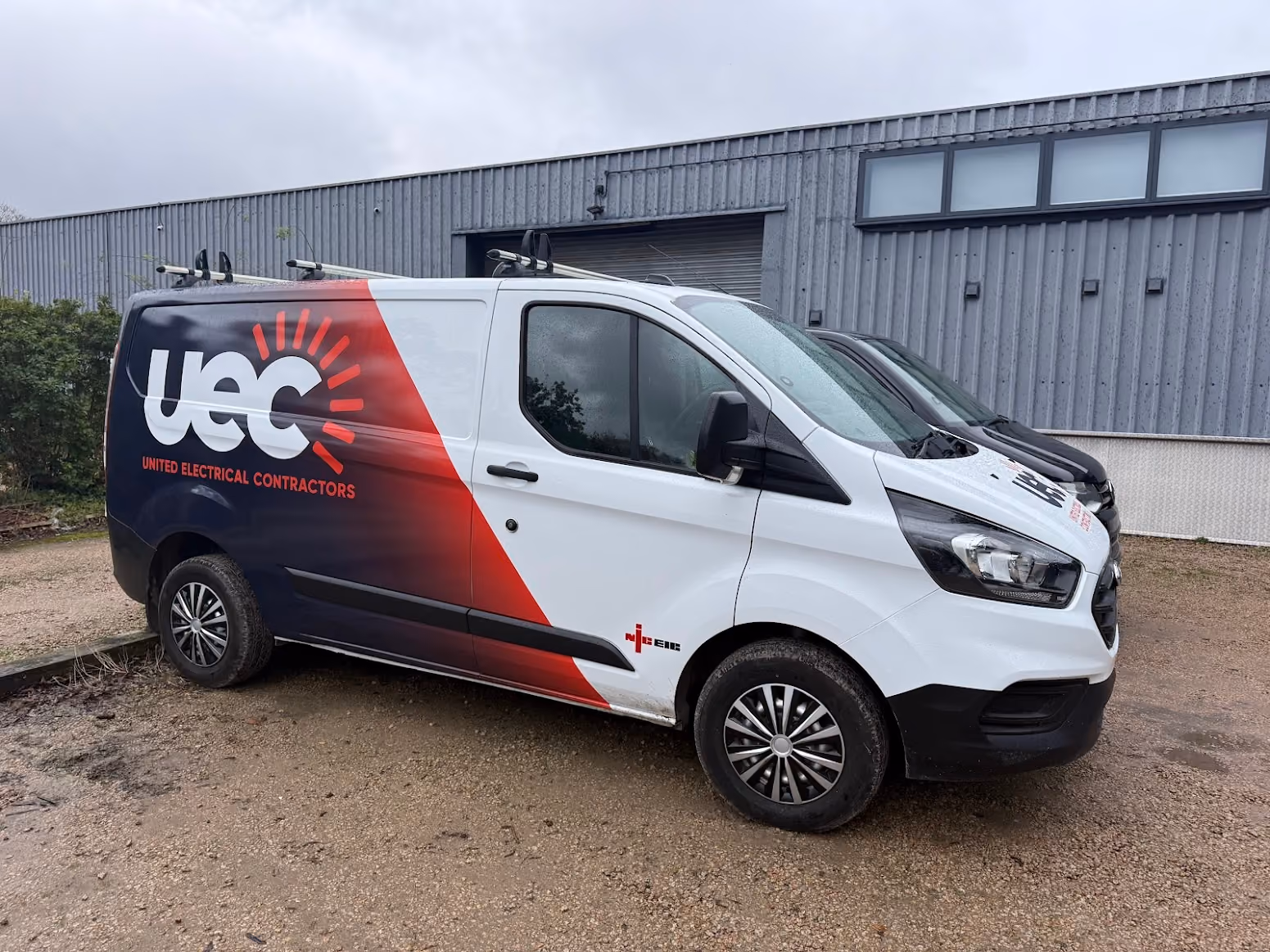 White and black commercial van parked outside a gray industrial building with the logo 'UEC' and 'United Electrical Contractors' on the side, featuring a red and black diagonal color split design.