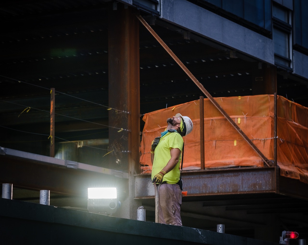 Construction worker in a yellow shirt and white hard hat leaning back while holding a shovel at a building site.