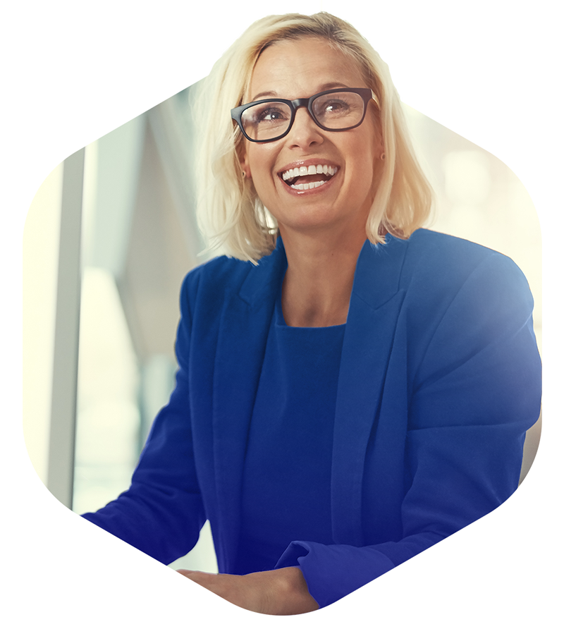 Smiling blonde woman wearing glasses and a blue blazer, sitting indoors near a window.