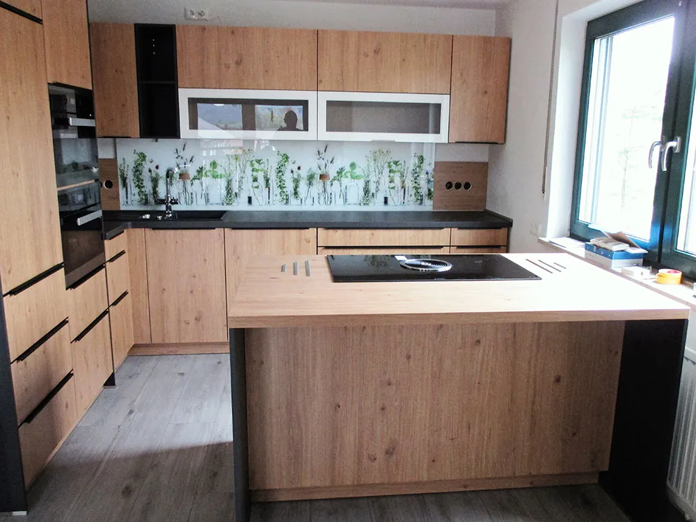 Moderne Küche mit Kochinsel, Holzfronten, schwarzen Arbeitsflächen und Rückwand mit Pflanzenmotiv.