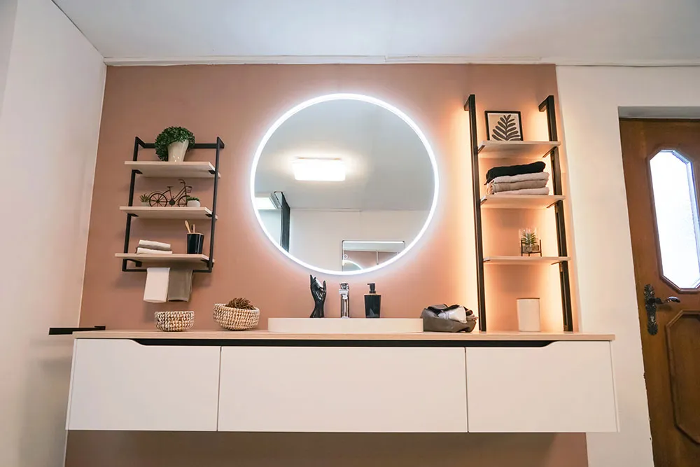 Modernes Badezimmer mit rundem, beleuchtetem Spiegel, offenen Regalen und weißem Waschbeckenunterschrank.