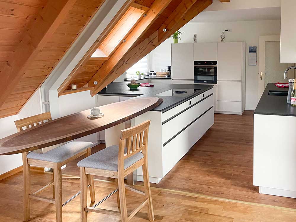 Moderne Dachgeschossküche mit weißen Fronten, Kochinsel, dunkler Arbeitsplatte und schräger Holzdecke.
