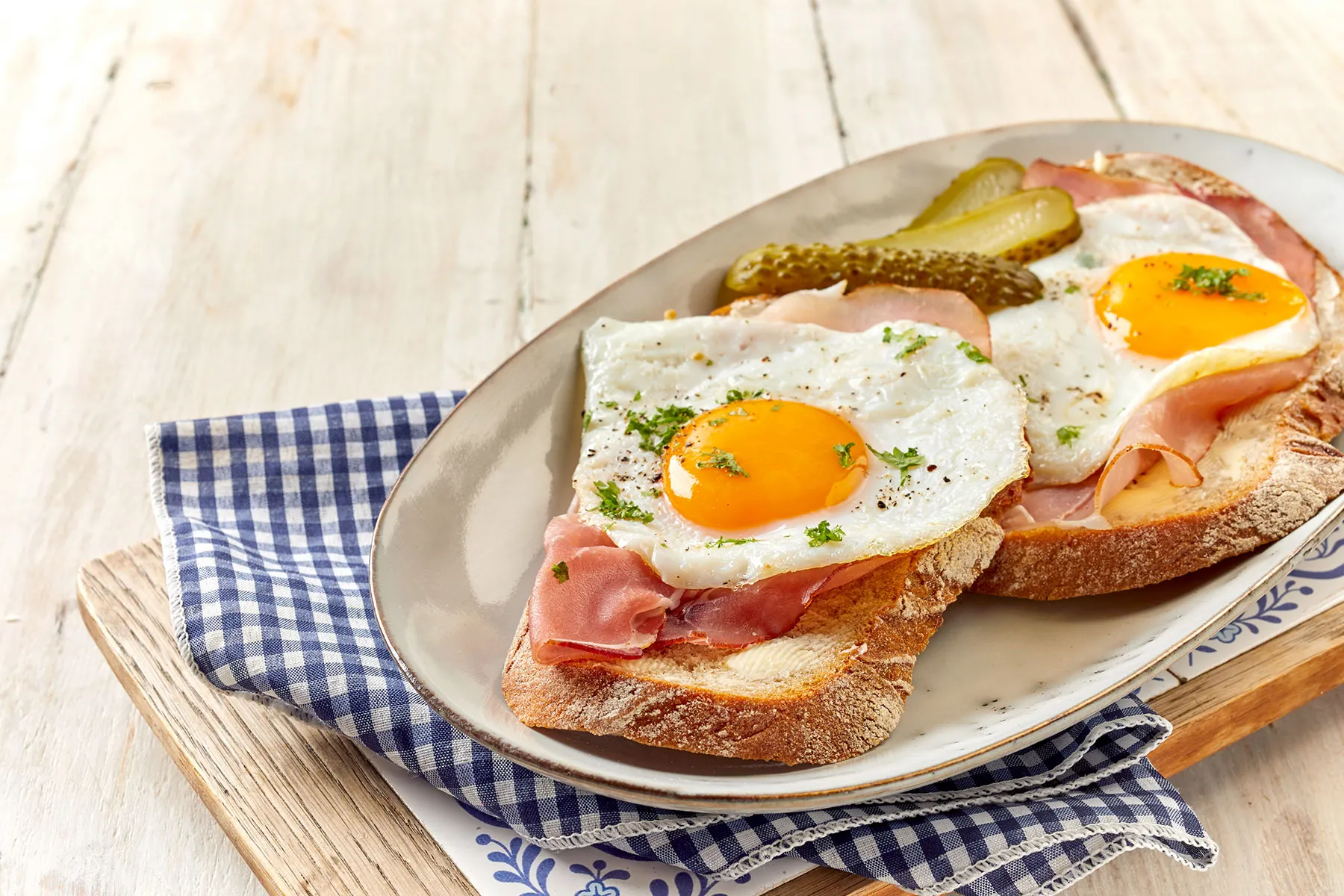 Ein Holzbrett mit einem Teller, auf dem zwei belegte Brotscheiben liegen, garniert mit Schinken, Spiegelei und Gewürzgurken.