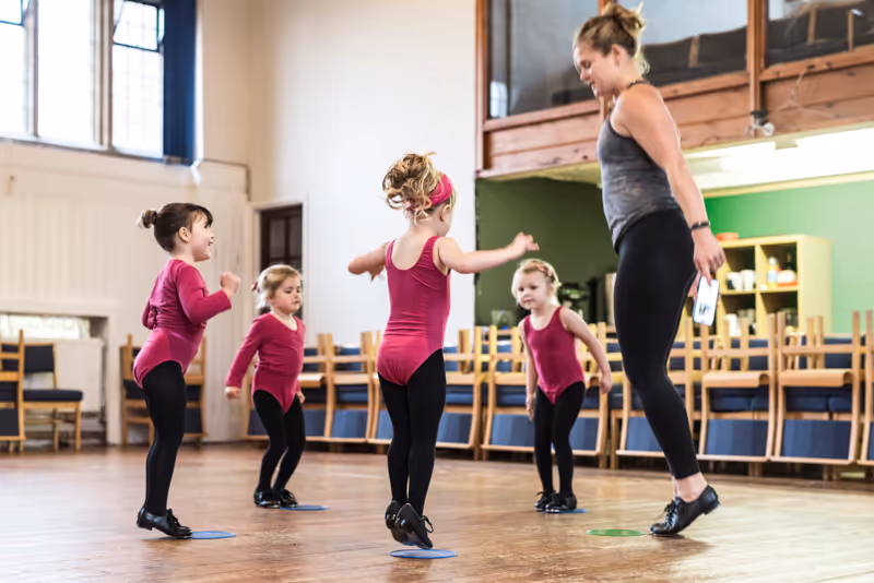 Dance instructor leading four young girls in matching pink tops and black leggings through a dance routine in a studio with wooden floor and chairs stacked in the background.