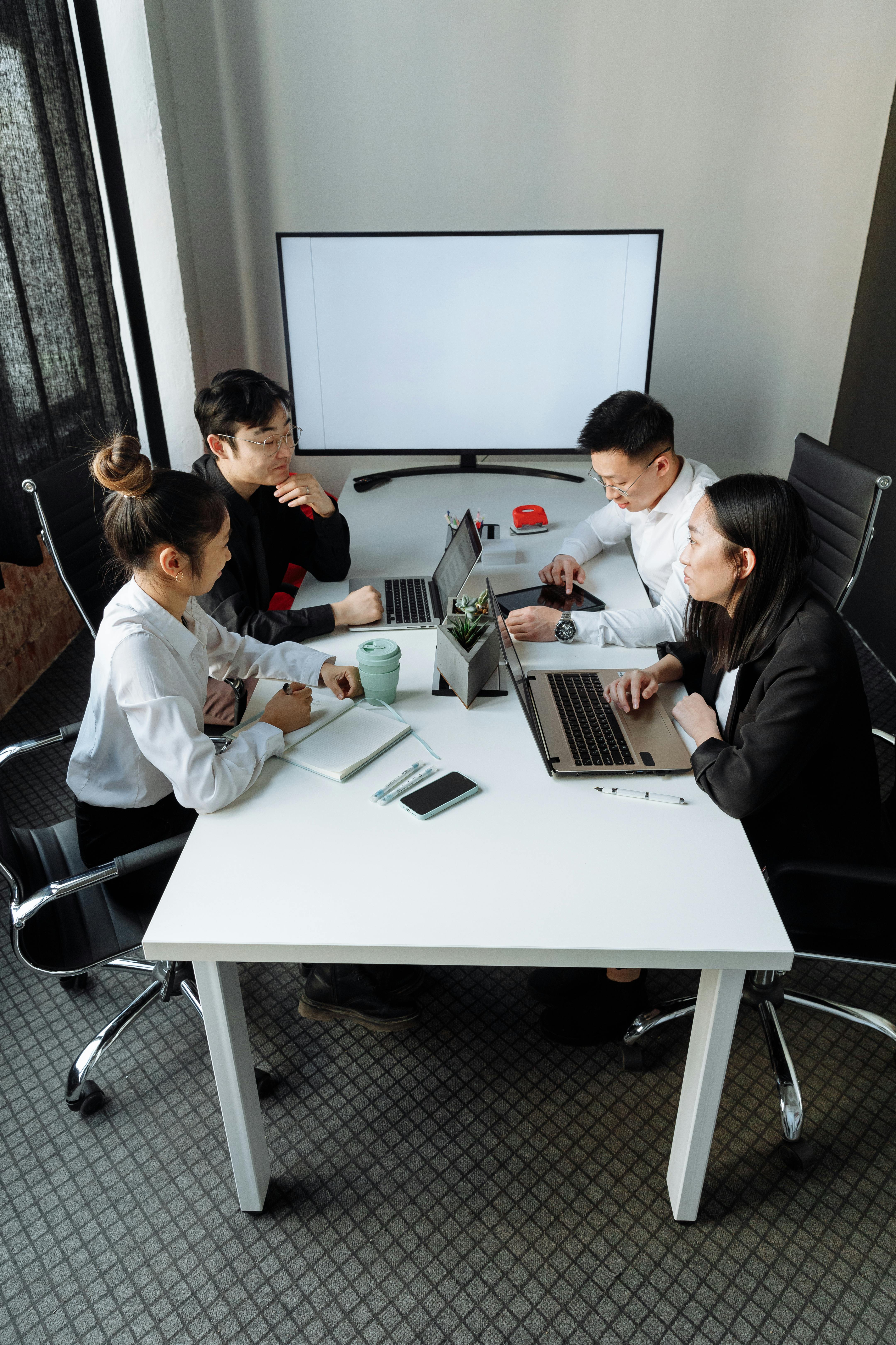 Four young professionals in a meeting room sitting around a white table with laptops, a notebook, and a large blank monitor on the wall.