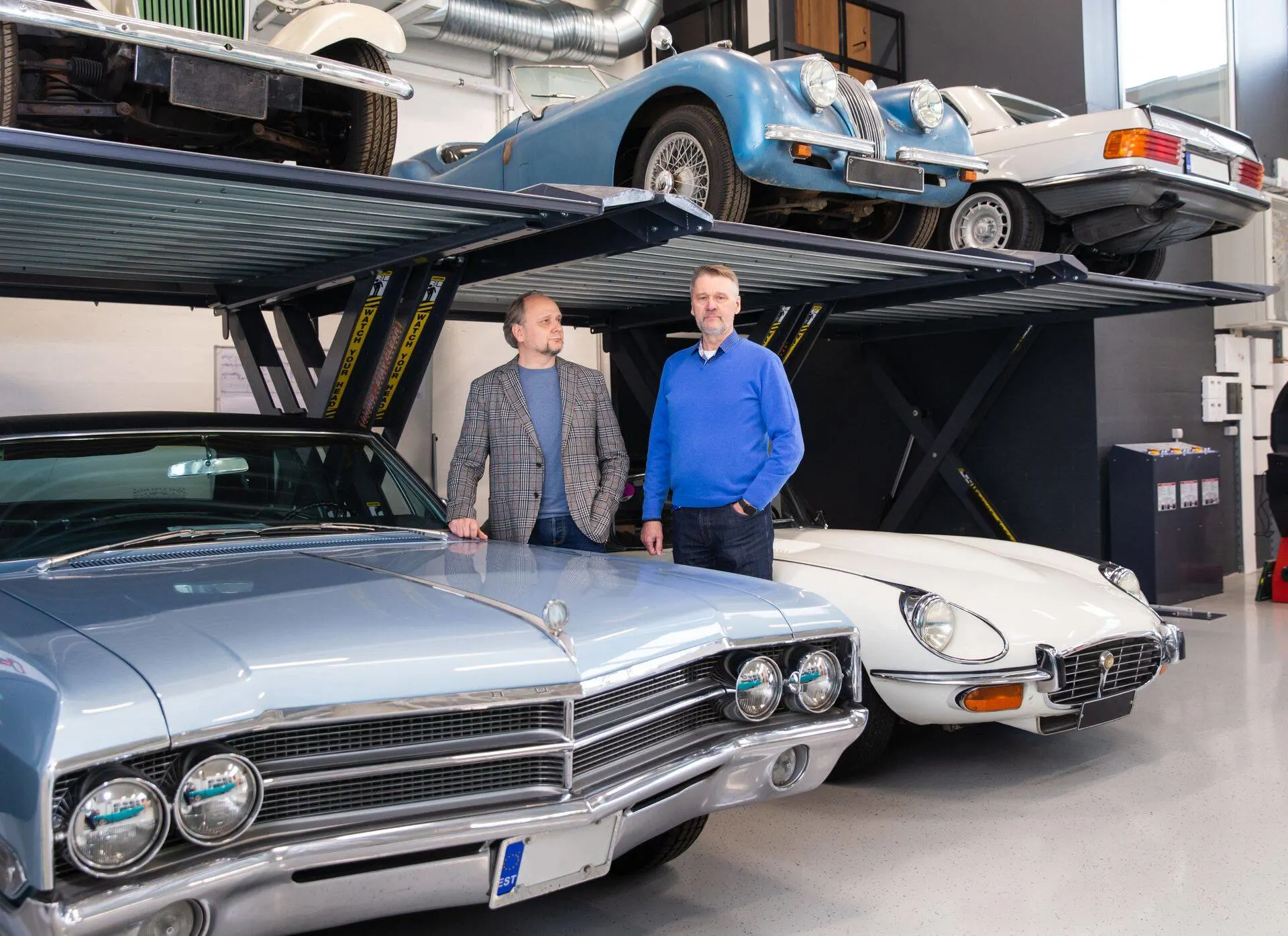 A picture of Peeter Neil Sarevet and Sulev Narusk standing in their workshop Benefit Auto Klassika between Buick Electra 225 and Jaguar E-type
